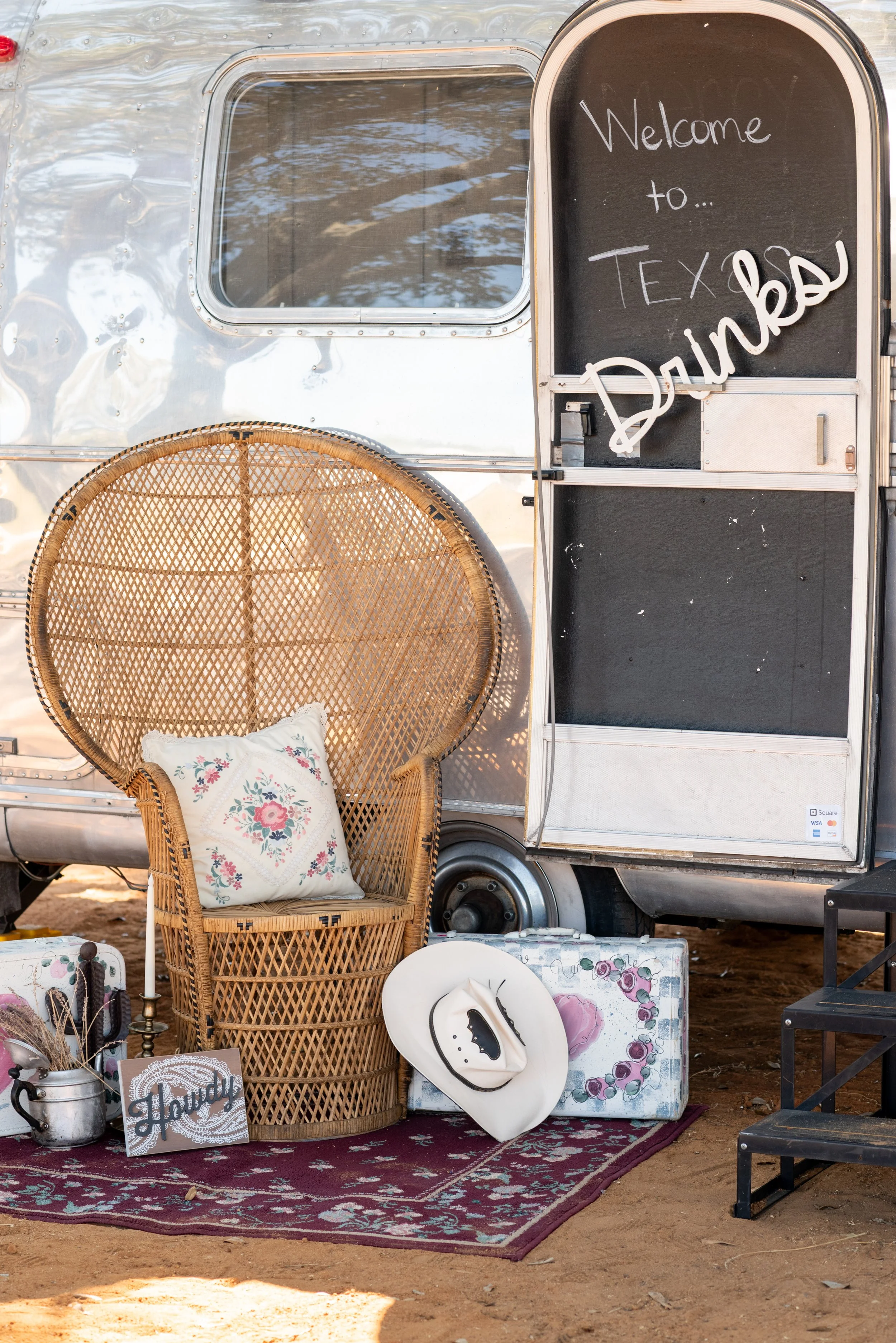 A vintage silver trailer with a window, a wicker peacock chair with a floral pillow, a white hat, and a decorative box on a red patterned rug. A chalkboard sign reads 'Welcome to... TEXAS DINKO'.