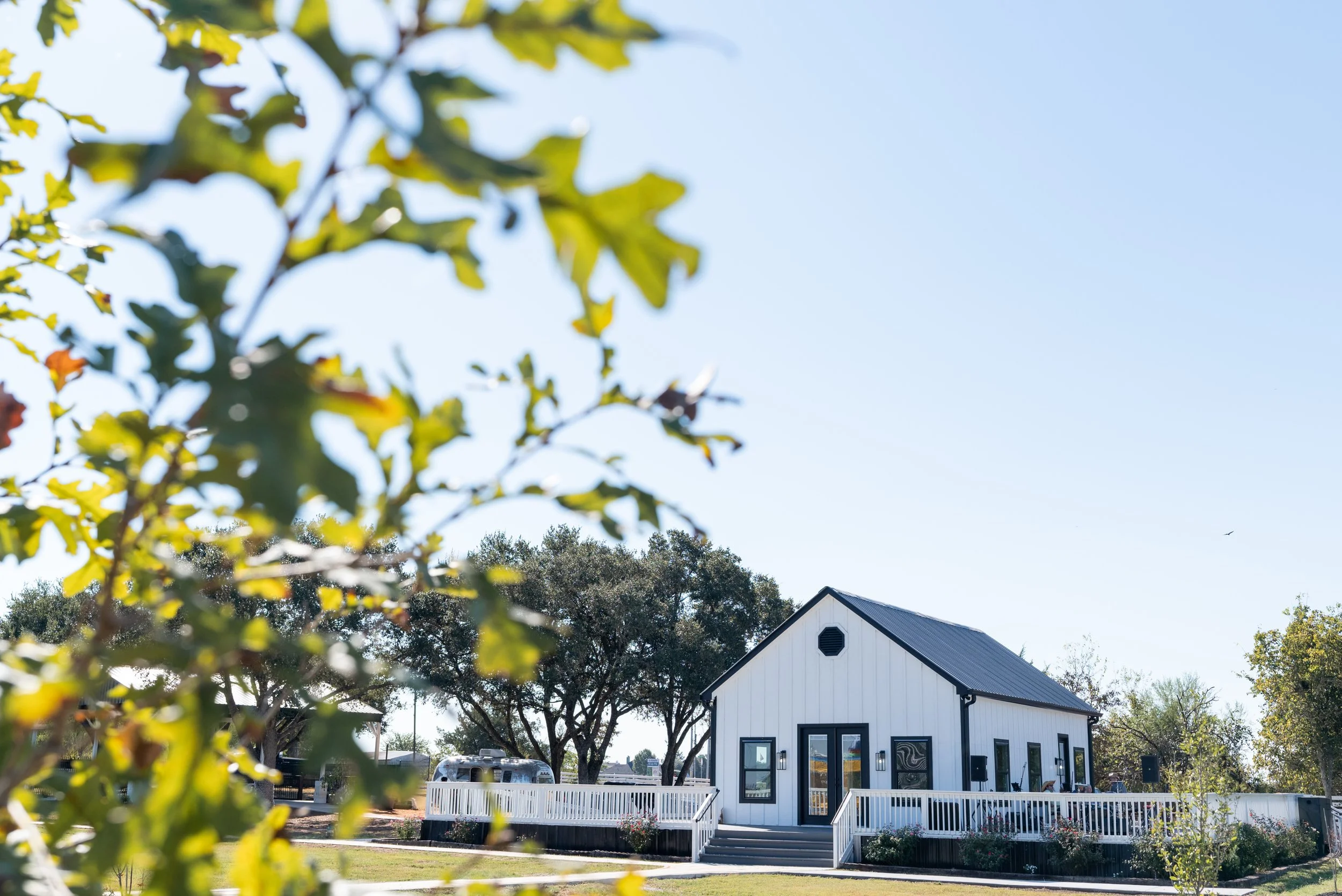 A white modern tiny house with black trim, large front door, and windows, surrounded by a white picket fence, with trees and a clear blue sky in the background.
