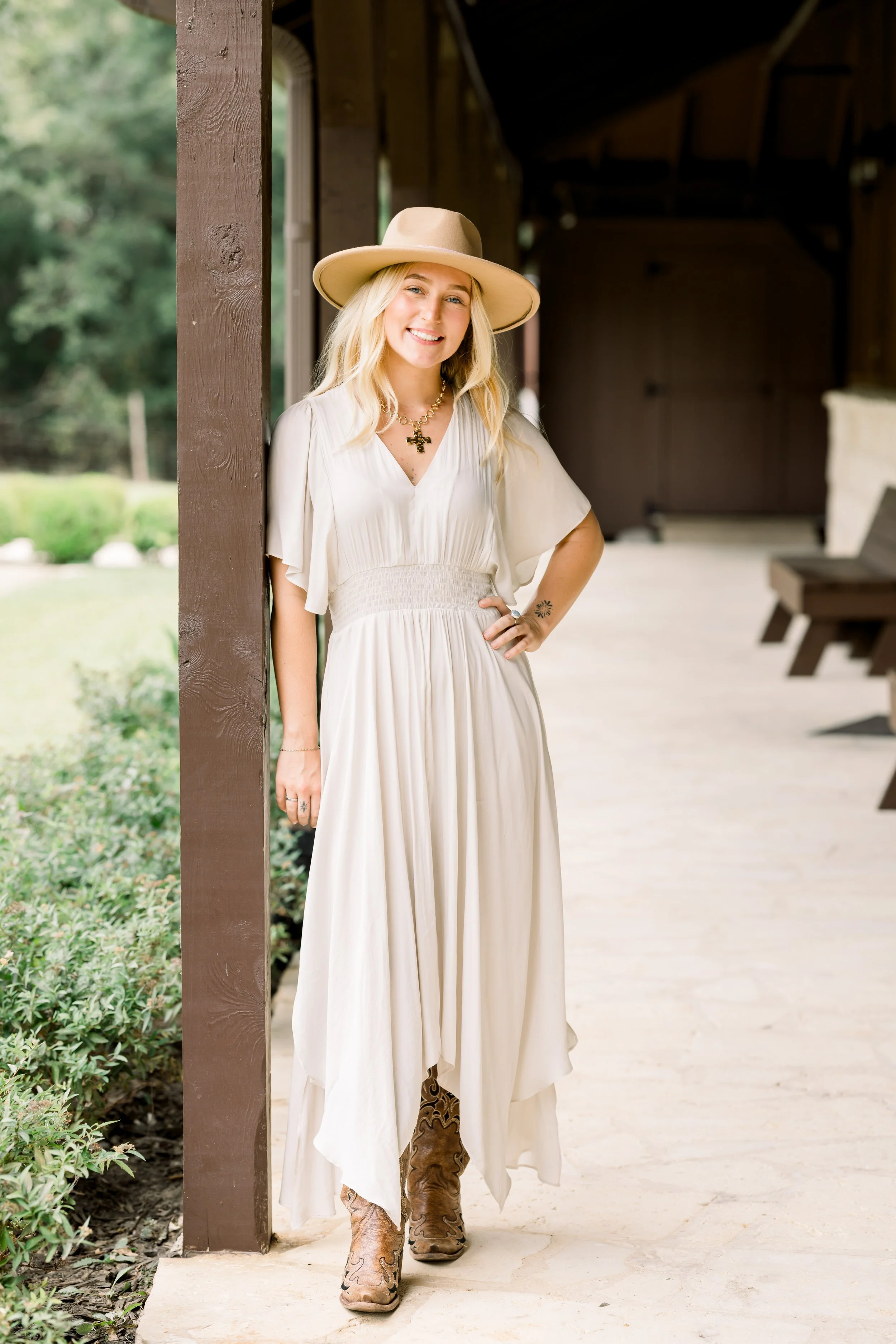 A woman with blonde hair wearing a cream-colored dress, a wide-brim hat, and cowboy boots, smiling and standing outdoors beside a wooden post.