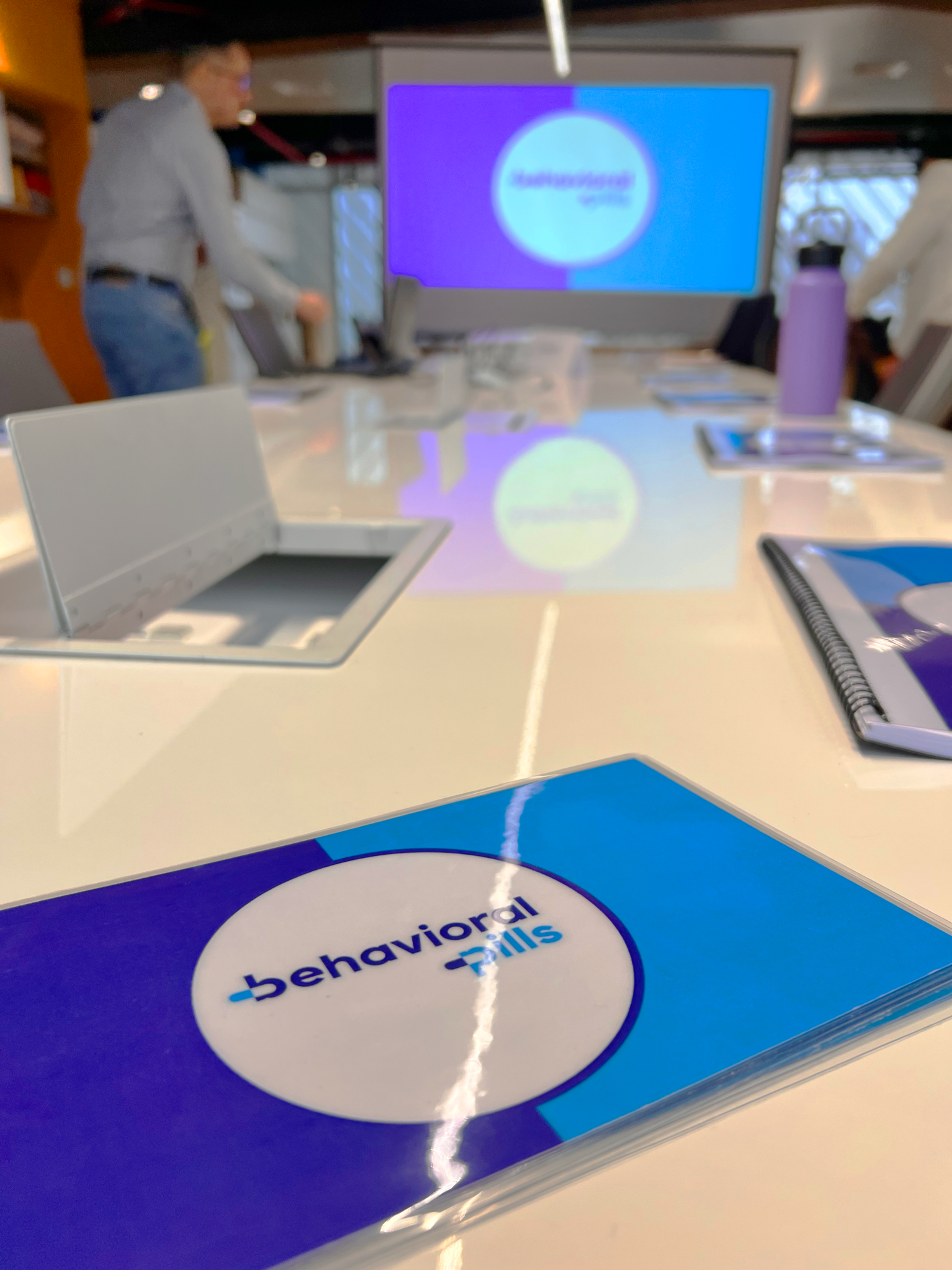 Una sala de reuniones con un libro titulado 'behavioral pills' sobre la mesa y una pantalla grande en la pared que muestra el logos y textos relacionados con una presentación o entrenamiento.
