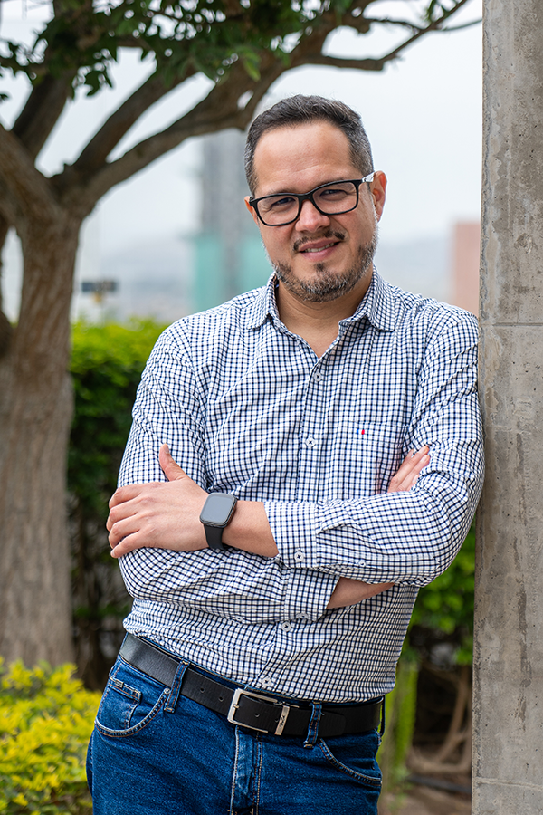 Hombre con gafas, barba, en una camisa de cuadros, de pie junto a una pared de concreto, en un entorno exterior con árboles y vegetación.