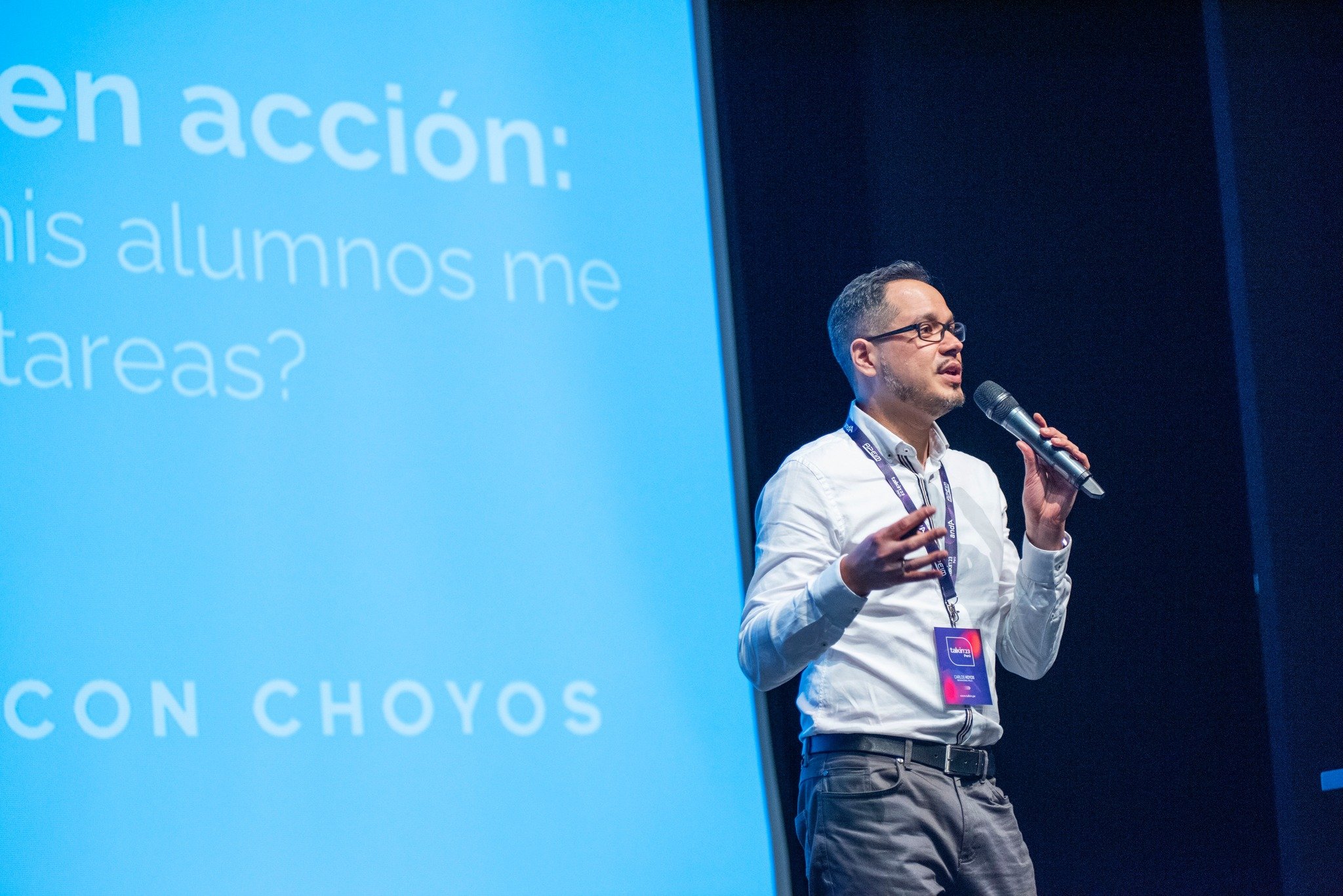 Hombre con gafas y camisa blanca dando una presentación con micrófono en una conferencia, con pantalla azul de fondo.