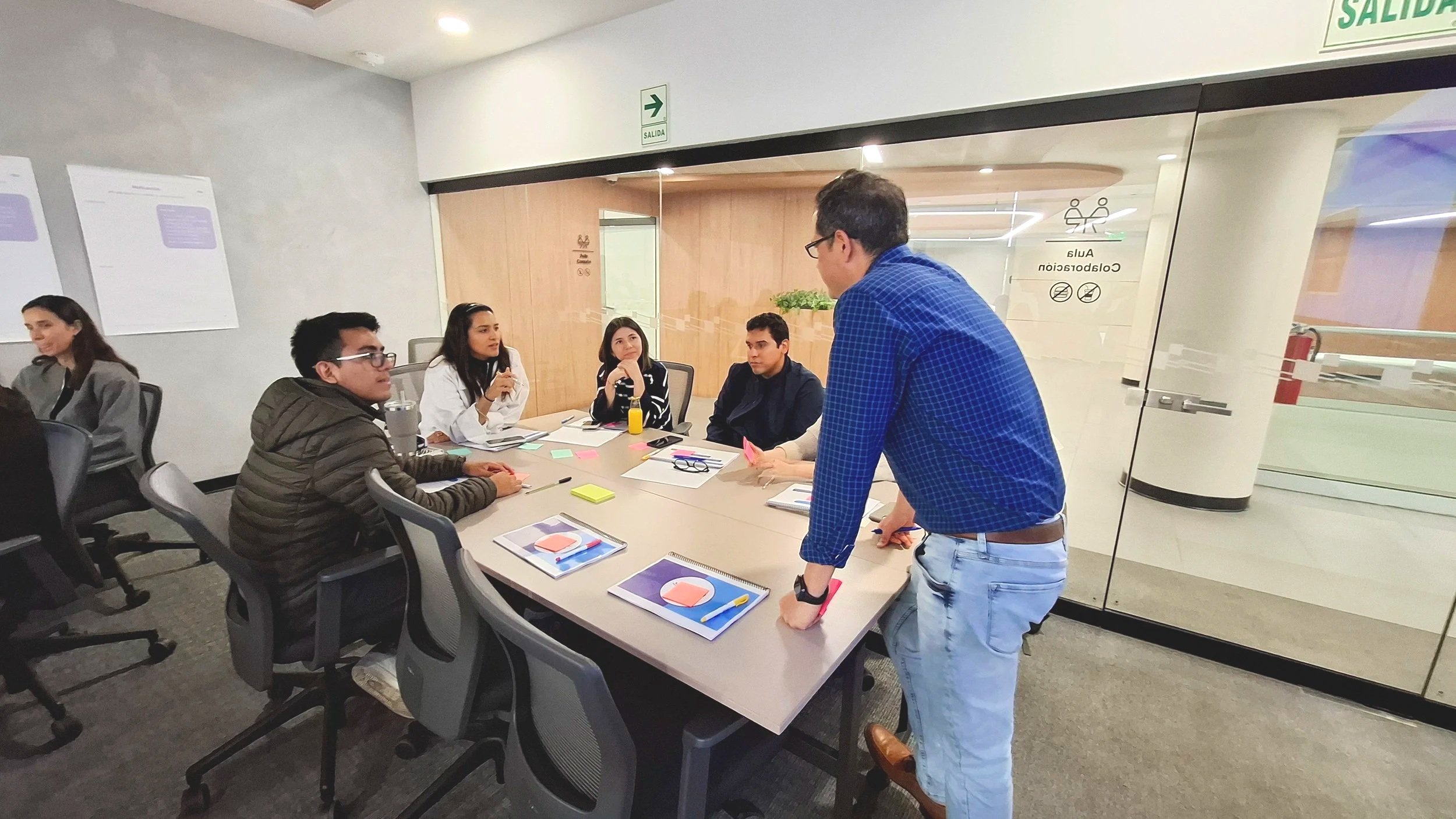 Grupo de personas en una reunión en una sala de conferencias, sentados en torno a una mesa con cuadernos y hojas, mientras un hombre de camiseta a cuadros y jeans se para y habla con ellos.