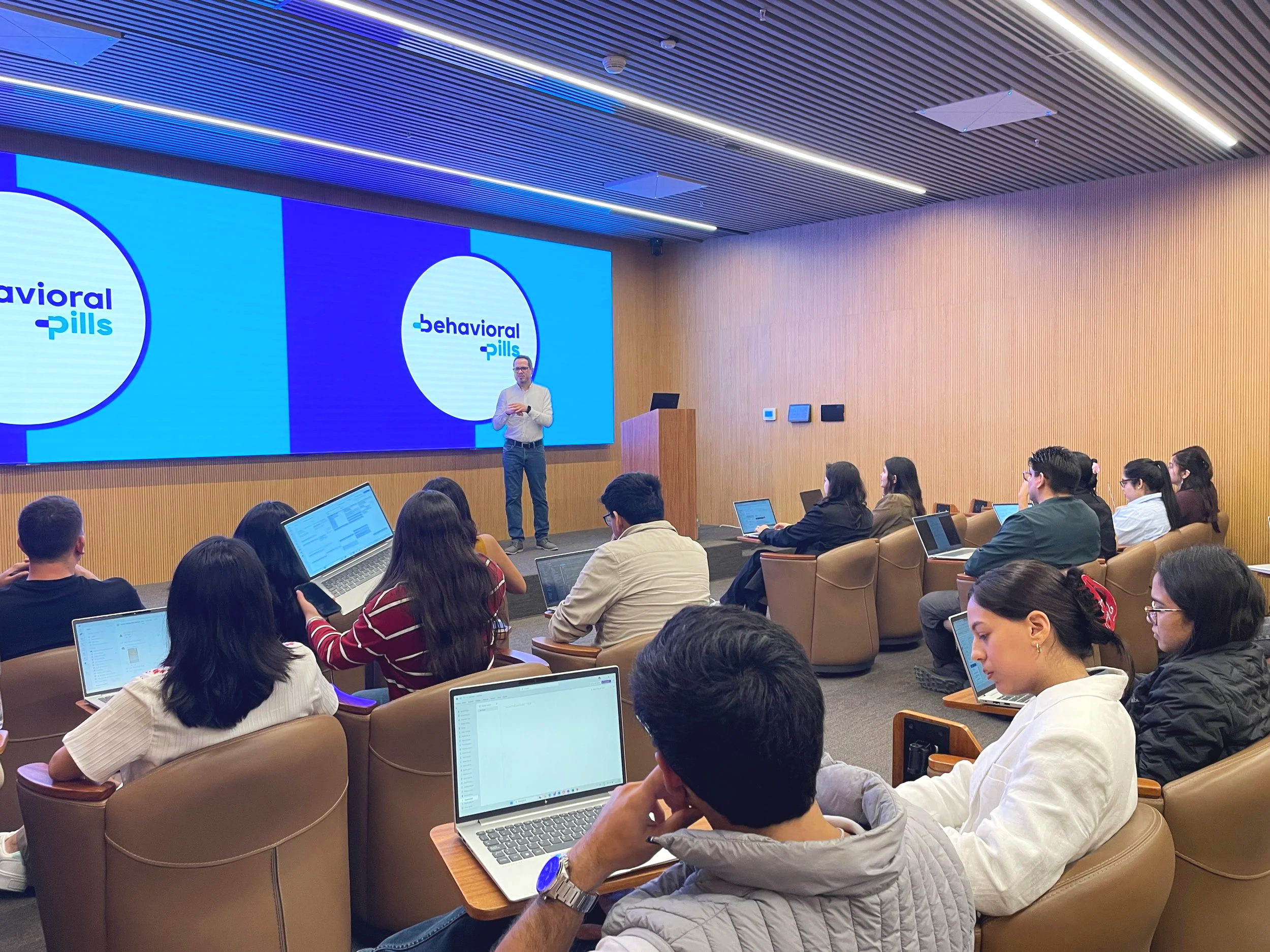 Salón de conferencias con un presentador en el escenario y audiencia usando laptops.
