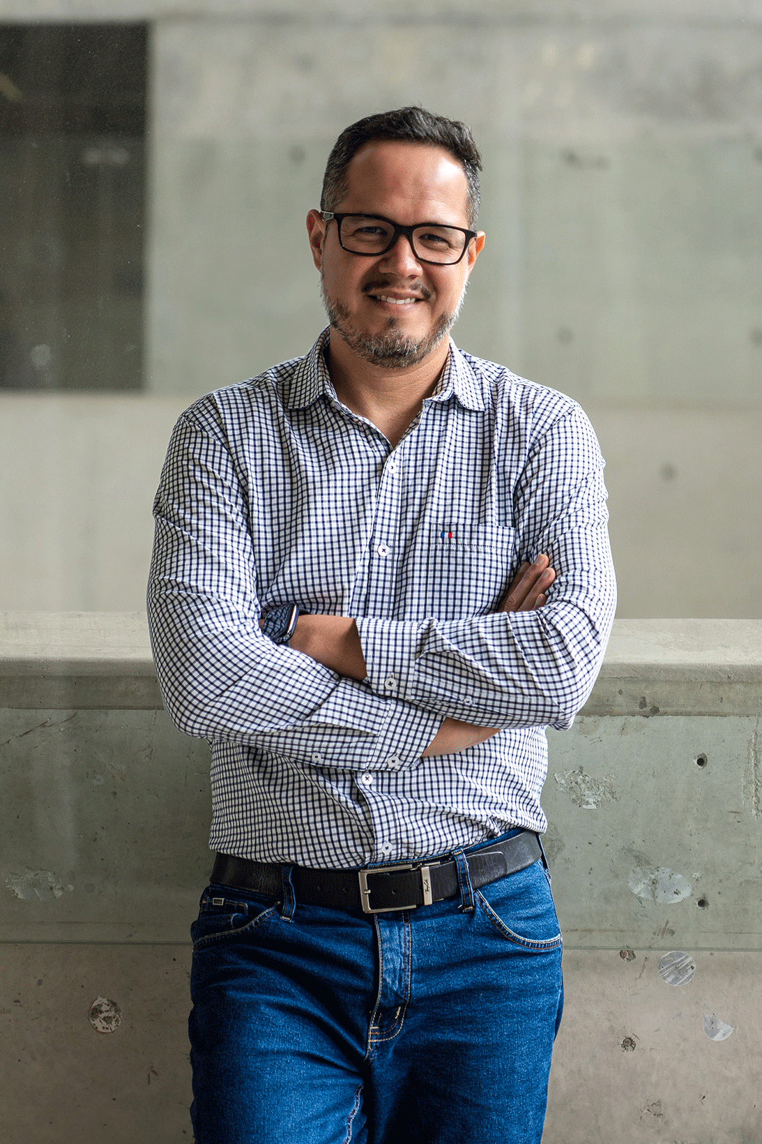 Hombre con gafas, barba, peinado corto, de camiseta de cuadros y jeans, cruzado de brazos, sonriendo frente a un fondo de concreto.