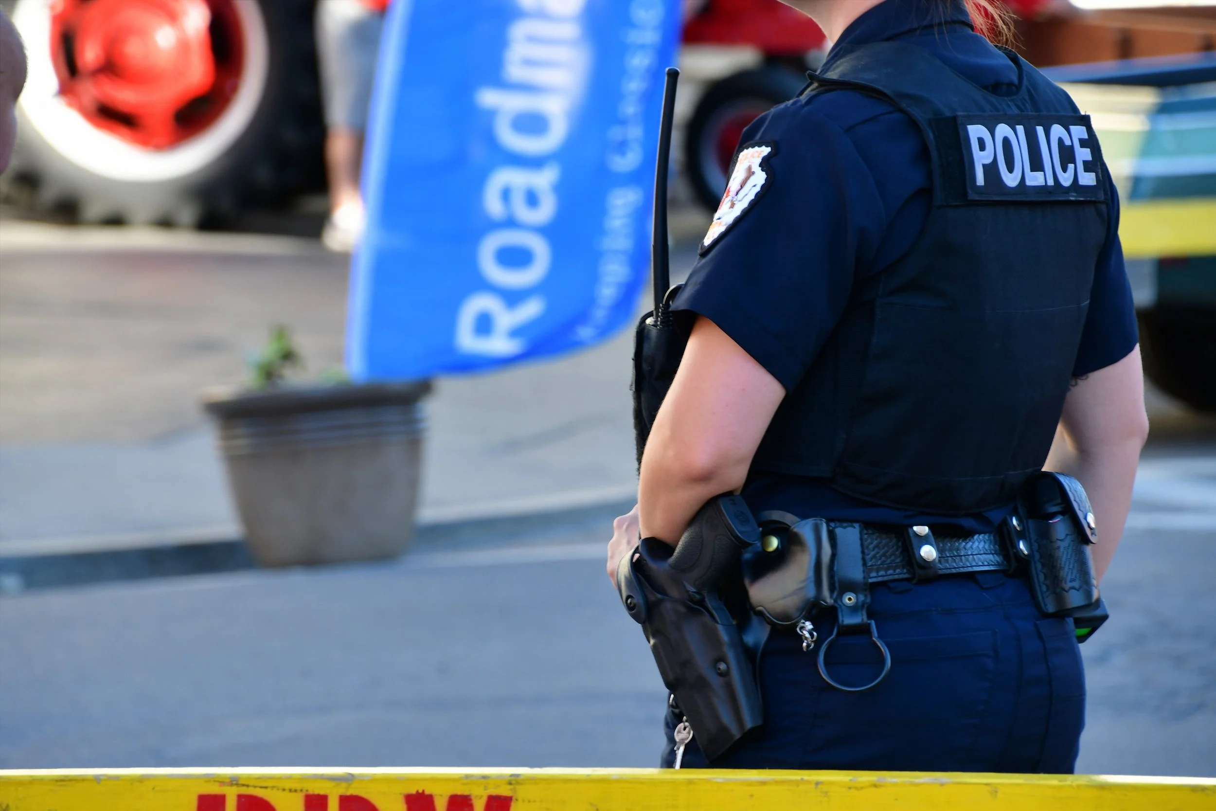 Close-up of a police officer wearing a dark blue uniform with a 'POLICE' patch on the back, standing near a yellow barrier with red text, with a blue flag or banner partially visible in the background.