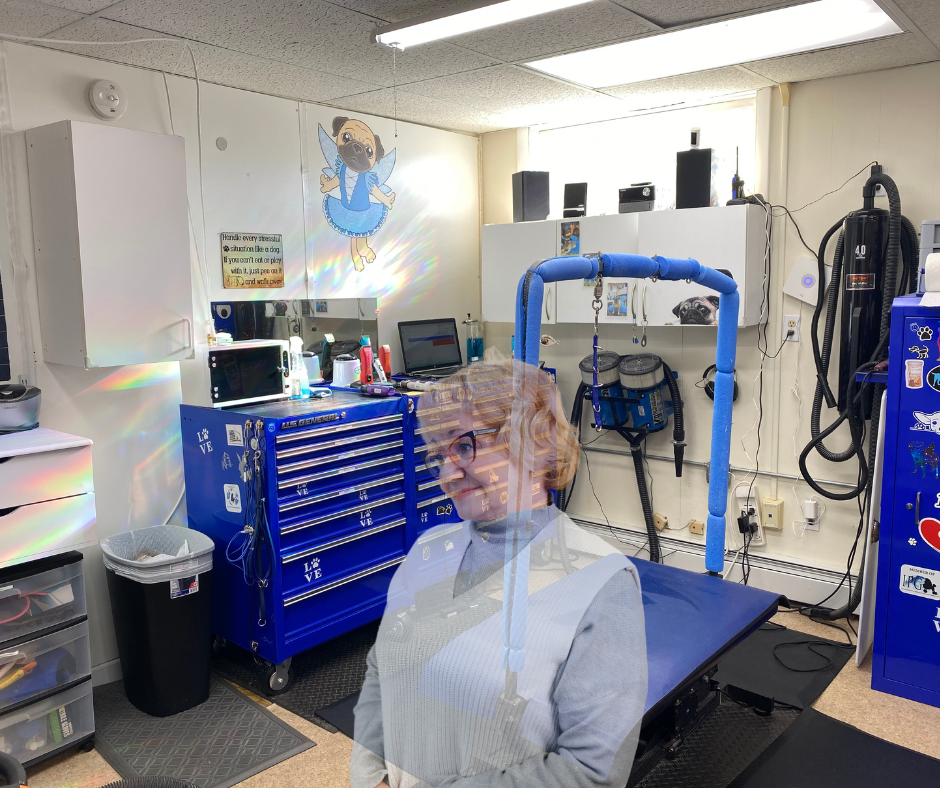 A woman with red hair and glasses standing in a pet grooming room with grooming equipment, a blue toolbox, and a cartoon dog poster on the wall.