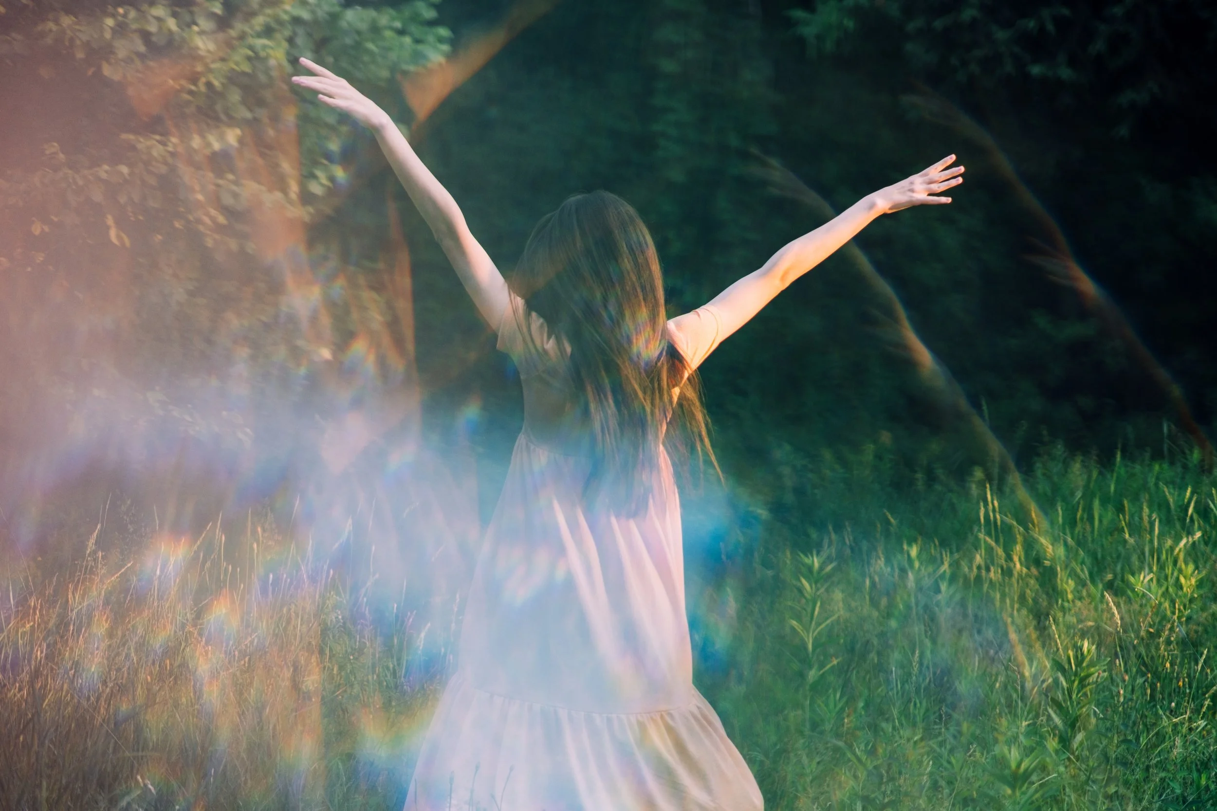 A girl with long hair in a light-colored dress standing in a grassy field with arms outstretched, surrounded by colorful light effects and lush green trees.