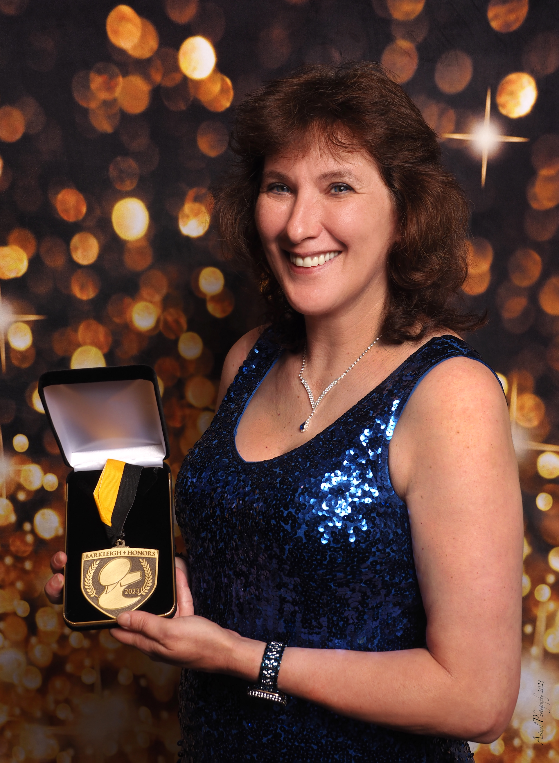 A woman in a blue sequined dress holding a gold medal in a black box, with a background of gold bokeh lights.