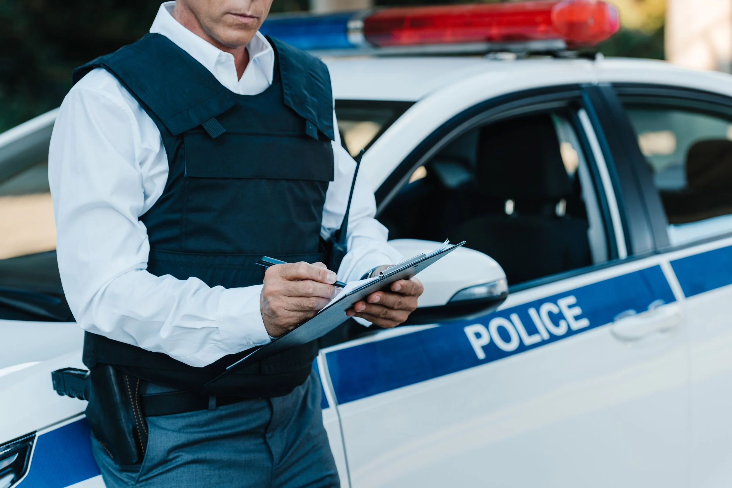 A police officer in a bulletproof vest writes in a notebook next to a police car with a red and blue light bar.