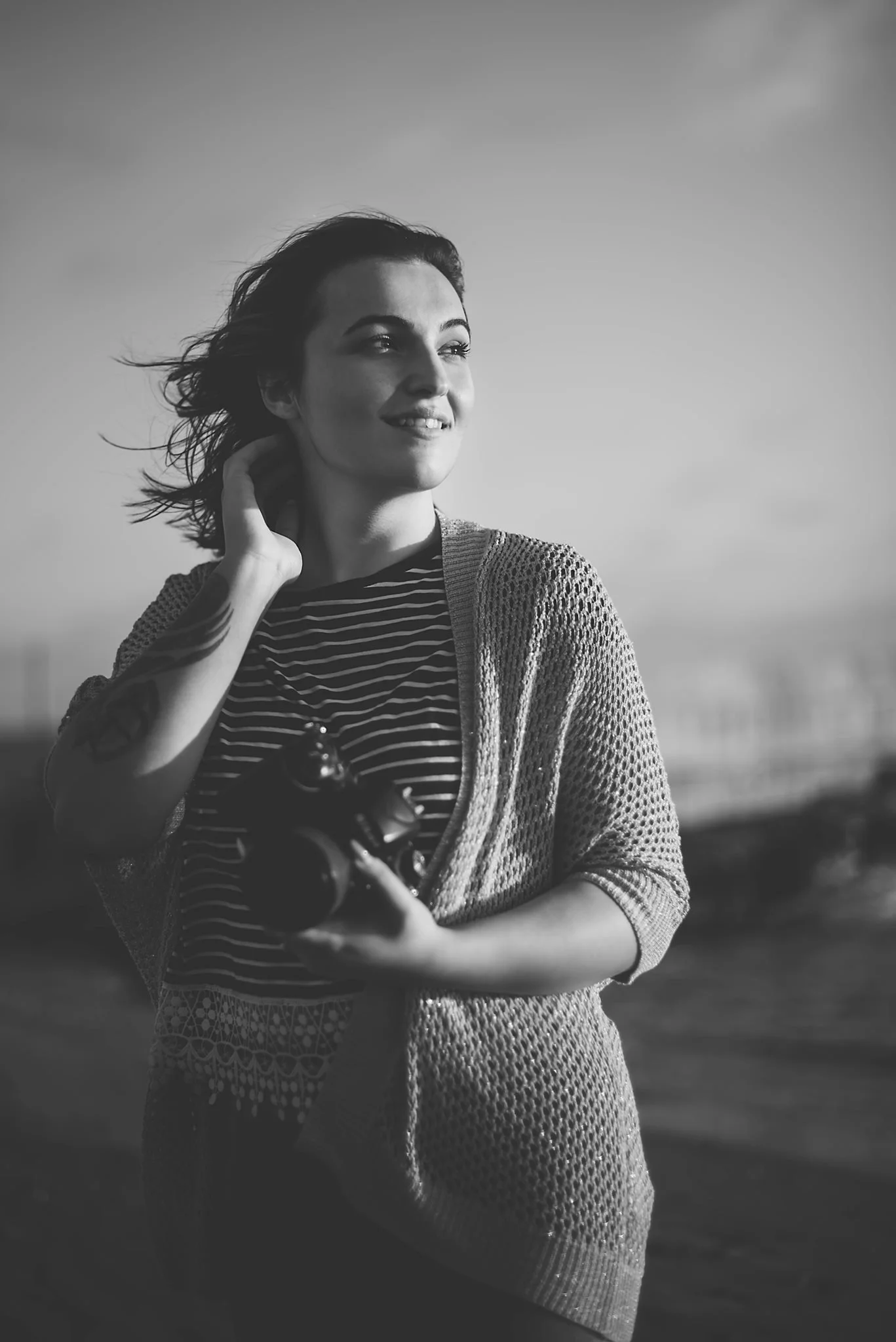 A woman standing outdoors holding a camera, smiling and looking away, with wind in her hair and dressed in a striped shirt and a knitted cardigan.