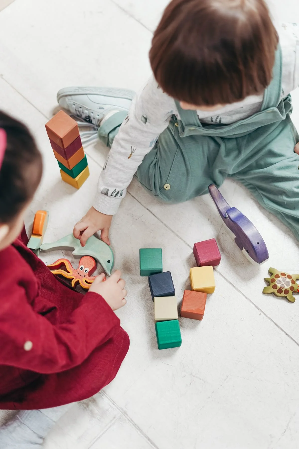 Two children playing with colorful wooden animal-shaped and block toys on a white tiled floor.
