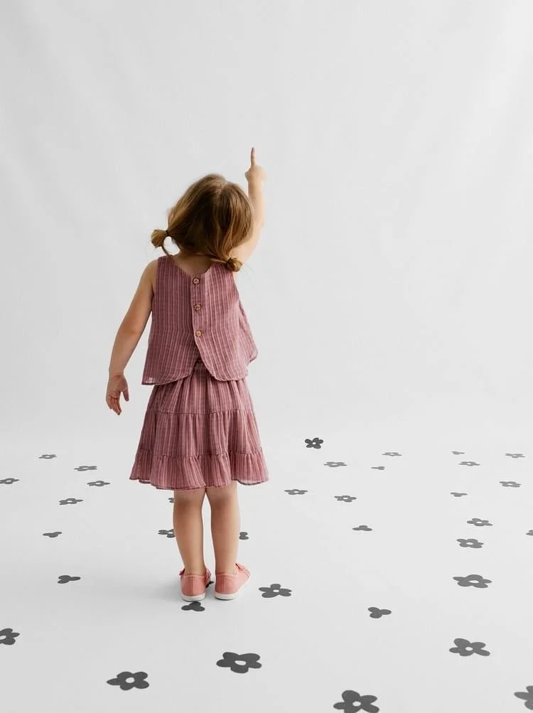 A young girl with brown hair in pigtails, wearing a pink dress and vest, pointing upward while standing on a white floor with scattered black flower shapes.