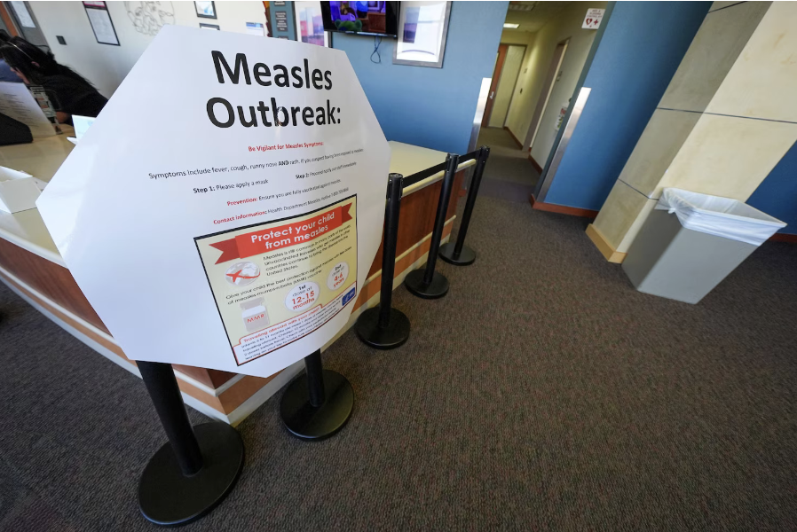 A large sign about measles outbreak with health advisories, placed on a stand in an indoor public space near a barrier and a trash bin.