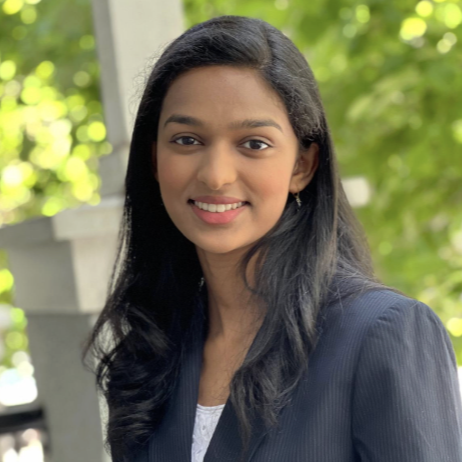 A young woman with long black hair smiling outdoors with green trees in the background
