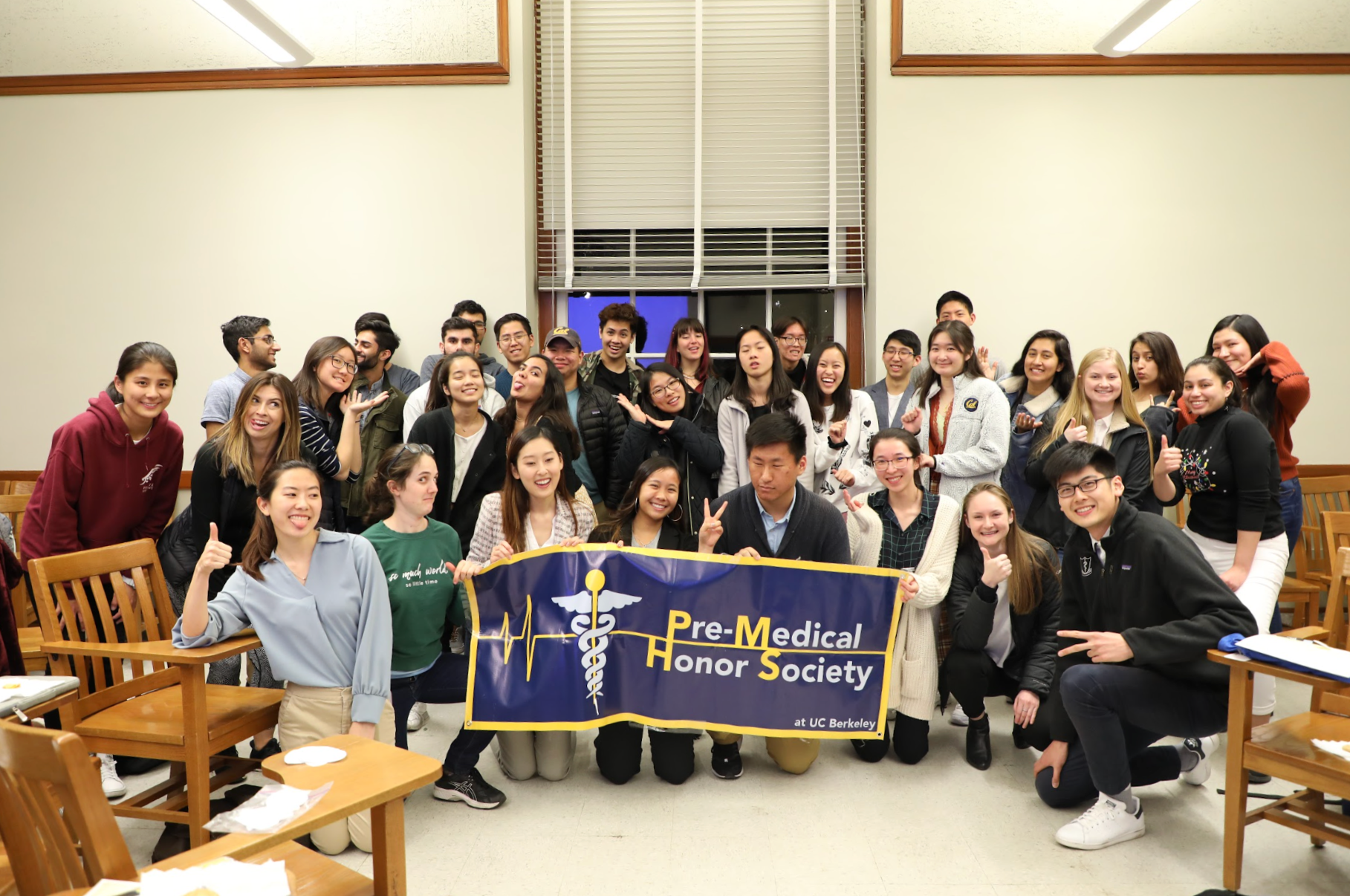 Group of diverse young people holding a banner that reads 'Pre-Medical Honor Society at UC Berkeley' in a room with wooden chairs and a large window.