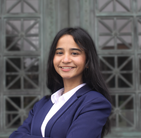 A smiling woman with long dark hair, wearing a navy blue blazer and white shirt, standing outdoors in front of a metal structure.