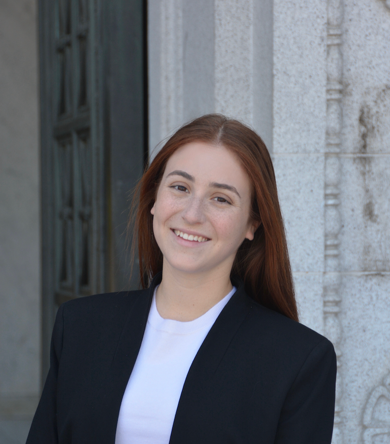 A young woman with red hair smiling, wearing a black blazer and white shirt, standing in front of a stone building with ornate detailing.