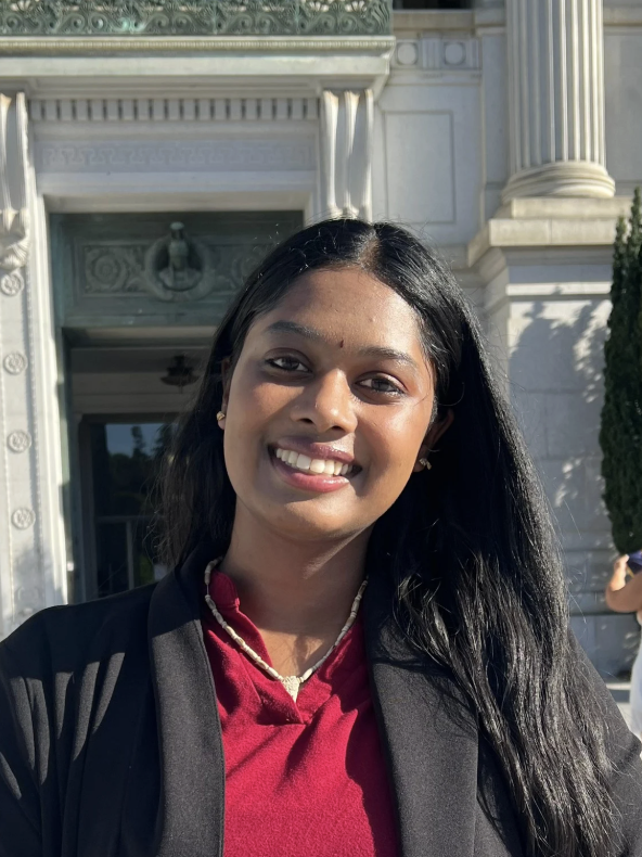 A young woman with long black hair and a bright smile, standing outside of a large ornate building with white stone columns and architectural details.
