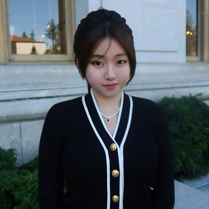 Young woman with short dark hair wearing a black cardigan with white trim, a pearl necklace, and a pendant. She is standing outdoors near a building with a window, greenery, and a clear sky in the background.