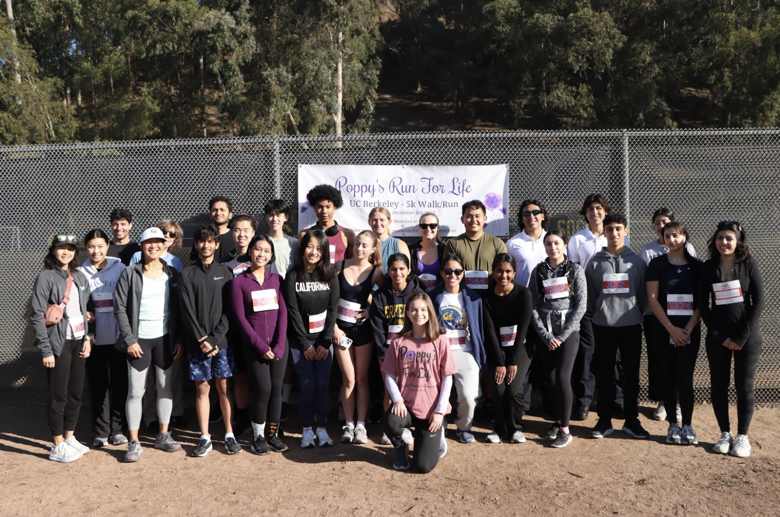 Group of diverse people posing for photo at a charity run event, with a banner in the background reading 'Poppy's Run For Life, UC Berkeley 5k Walk/Run'.