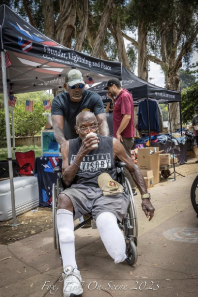 An elderly man in a wheelchair drinking from a cup, with two men standing behind him, at an outdoor event with tents and American flags.