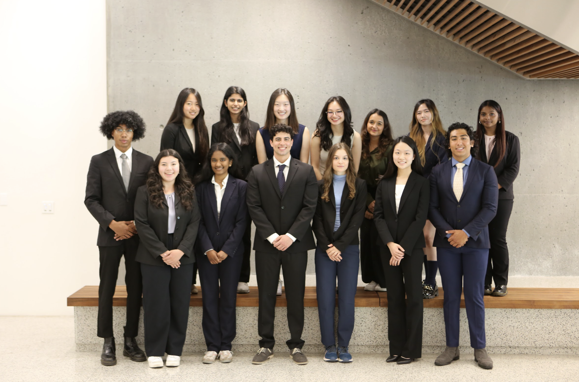 A diverse group of fifteen young professionals, both men and women, dressed in formal business attire, standing on a wooden platform in front of a gray concrete wall indoors, smiling at the camera.