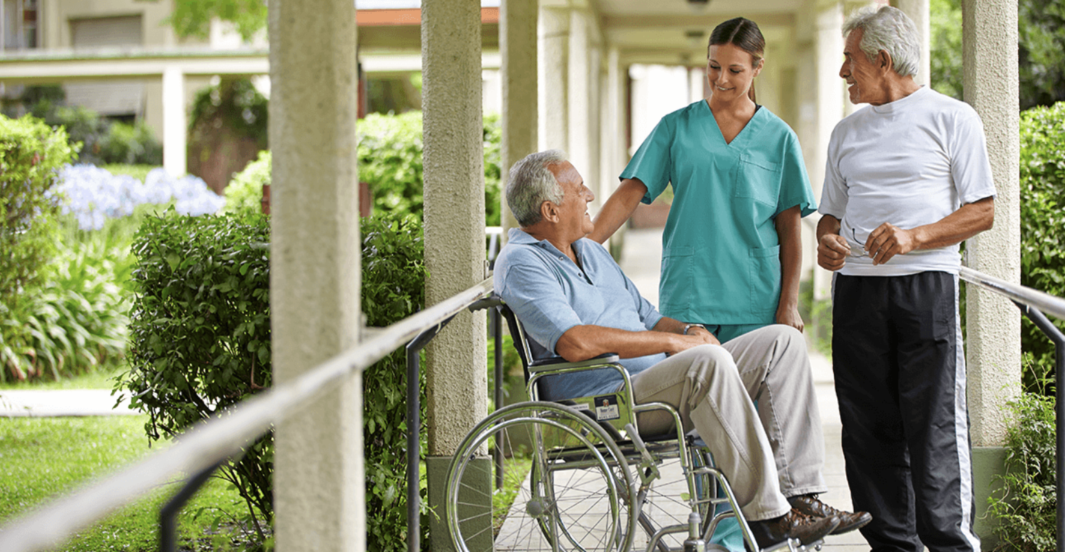 A healthcare worker smiling and talking with an elderly man in a wheelchair outdoors, with a second elderly man standing nearby in a garden setting.