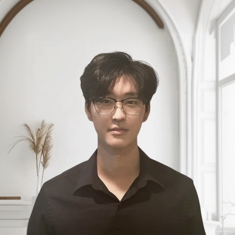 A young man with black hair, wearing glasses and a black shirt, standing in front of a white wall with a window and decorative grasses in the background.