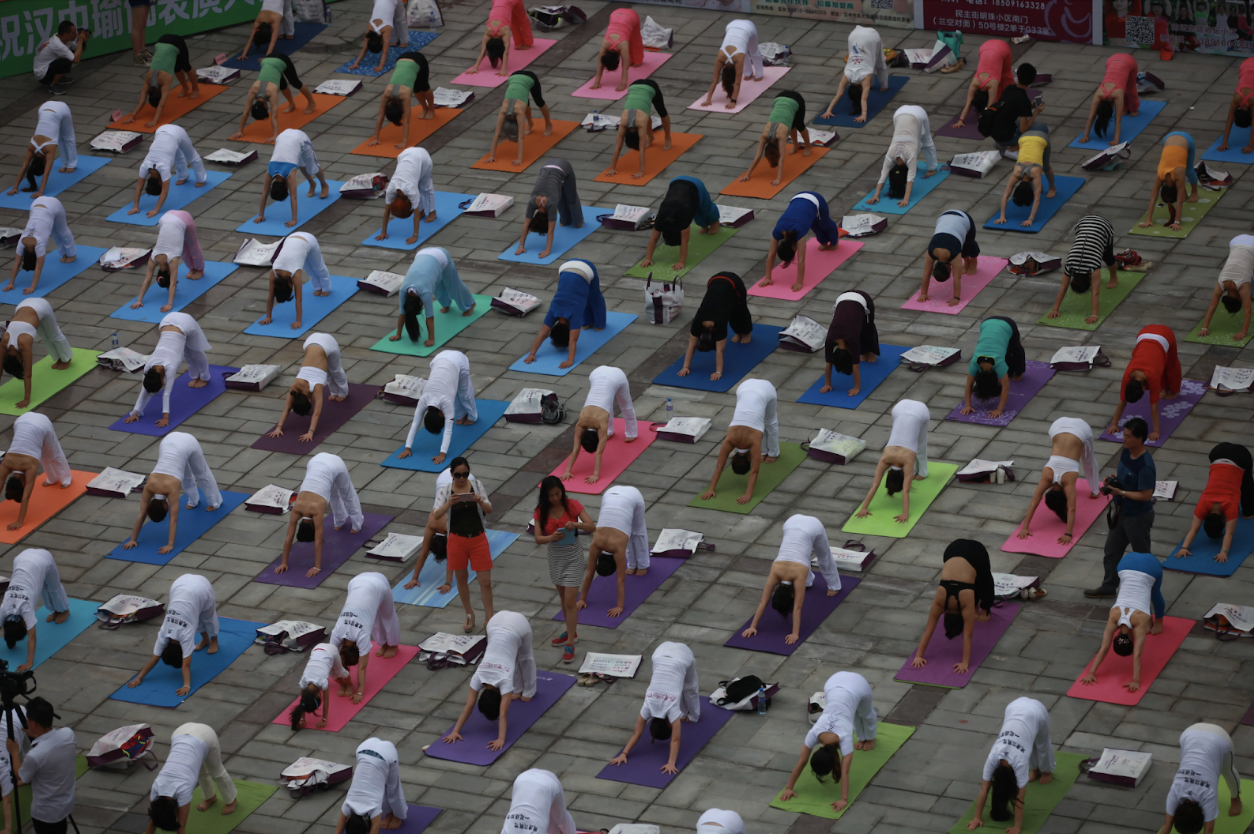 A large group of people practicing yoga outdoors on colorful mats arranged in neat rows on a stone paved surface. Everyone is in a downward dog pose, with some individuals reading or looking at books placed nearby.