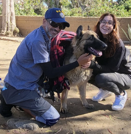 Two people, a man and a woman, are kneeling outdoors with a large German Shepherd dog between them. The man is wearing a blue cap, sunglasses, and a blue shirt, while the woman has long hair, glasses, and is dressed casually. They are smiling and appear happy.