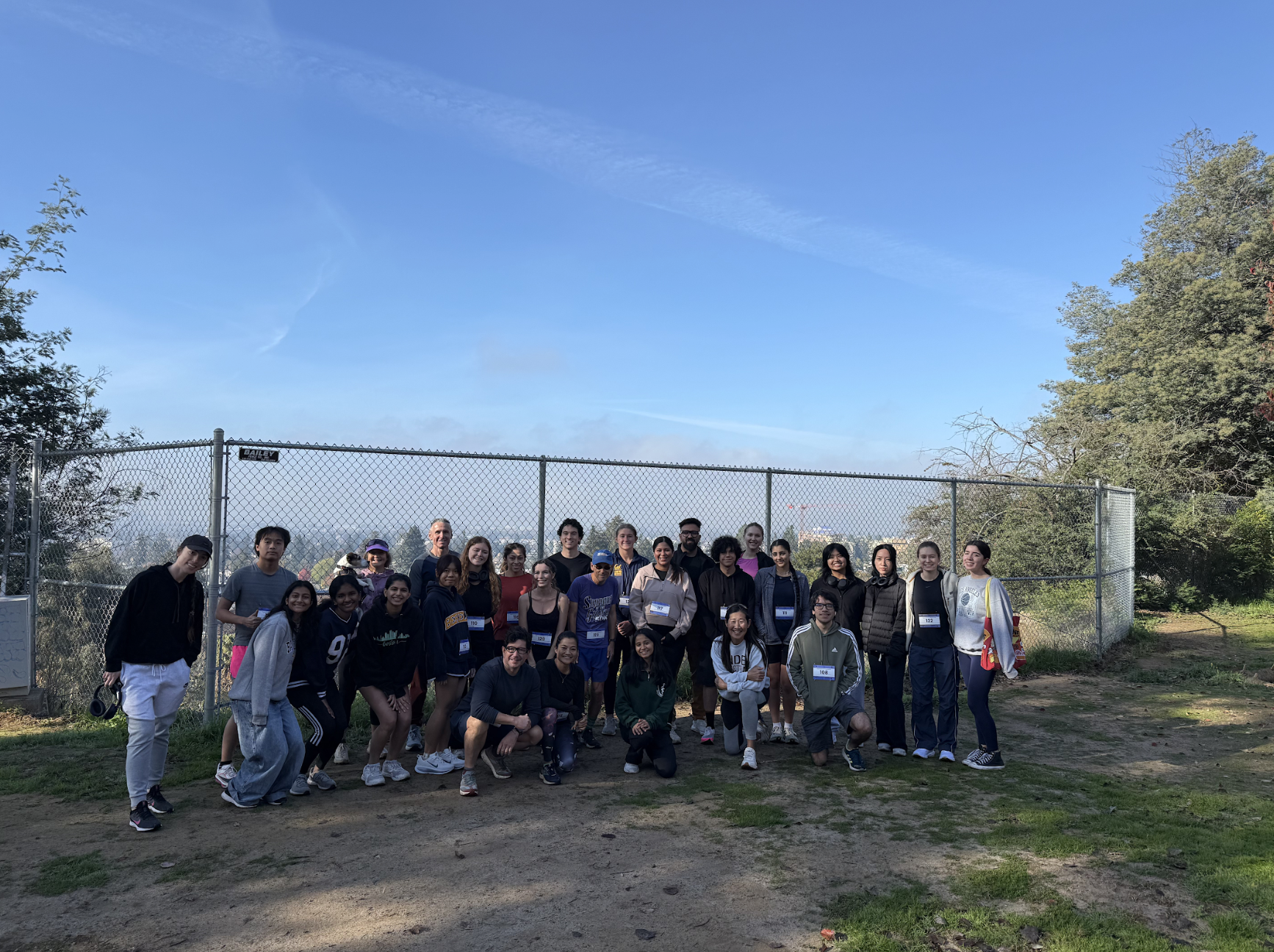 Group of diverse people posing outdoors in front of a chain-link fence beneath a clear blue sky with scattered clouds.