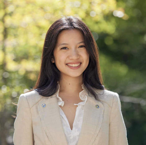 A woman with long dark hair smiling outdoors with greenery in the background.