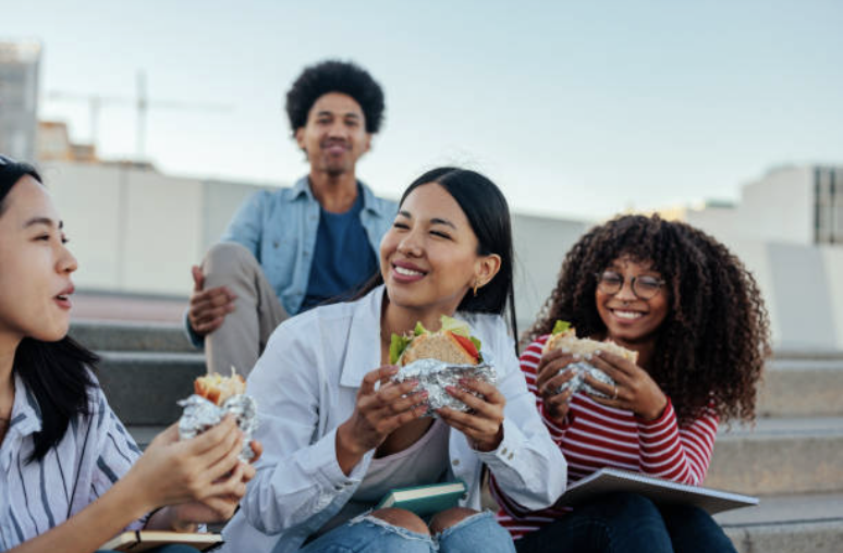 A group of four young friends sitting on outdoor steps, enjoying sandwiches and smiling.