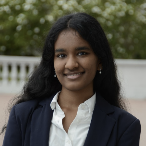 A young woman with long black hair smiling outdoors, wearing a navy blazer, white shirt, and pearl earrings.