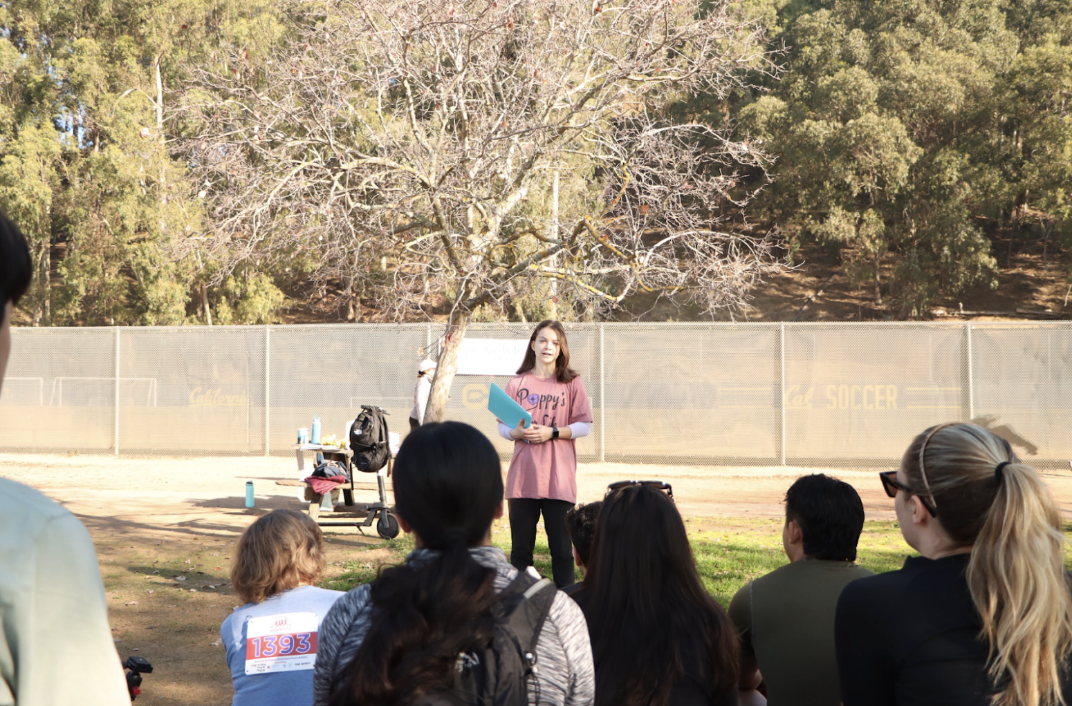 A young woman standing outdoors near a leafless tree, speaking to a group of people seated on the grass or benches, with a sports field and trees in the background.
