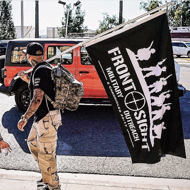 A man walking on sidewalk carrying a large black flag with white text and silhouettes of soldiers, with a red and black Jeep parked behind him.