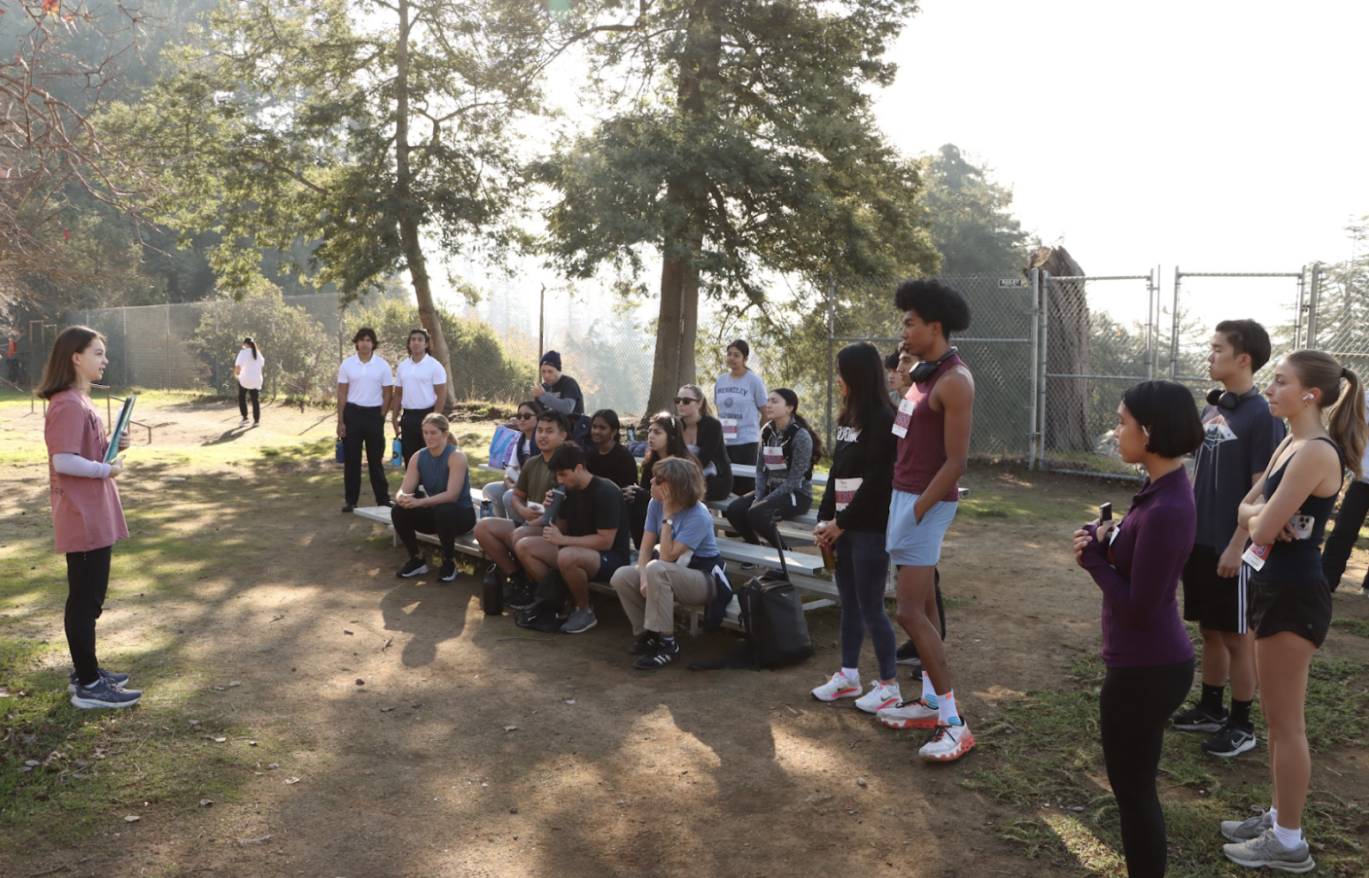 A group of young people attending a class outdoors in a park, sitting on benches or standing, while a woman in pink teaches or speaks to them. The setting includes trees, grass, a dirt ground, and a chain-link fence in the background.
