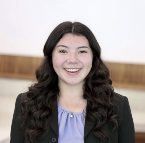 A young woman with long dark wavy hair, wearing a black blazer over a light purple blouse, smiling in an indoor setting.