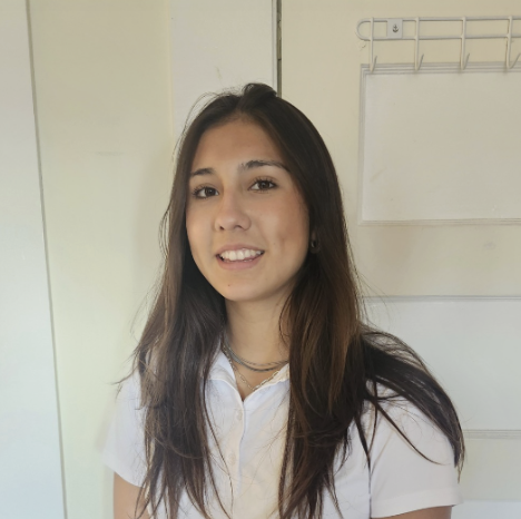 Young woman with long dark hair wearing a white polo shirt, standing indoors in front of a white wall with a metal rack.