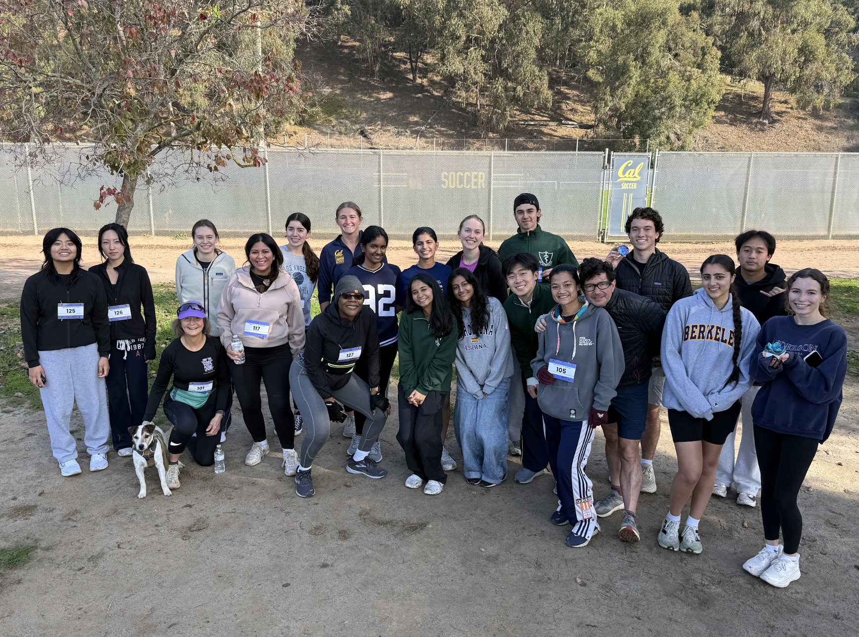 Group of people, mostly young adults, standing outdoors on a dirt path in front of a fence with a sign that says 'Cal SOCCER'. Some are wearing workout clothes and tags with numbers, and there is a dog on a leash near a woman kneeling in front.