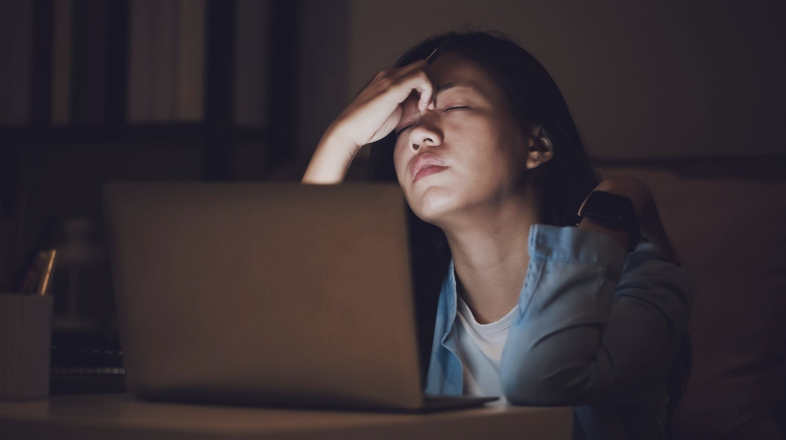 A woman sitting at a desk with a laptop, holding her head in frustration or fatigue in a dimly lit room.