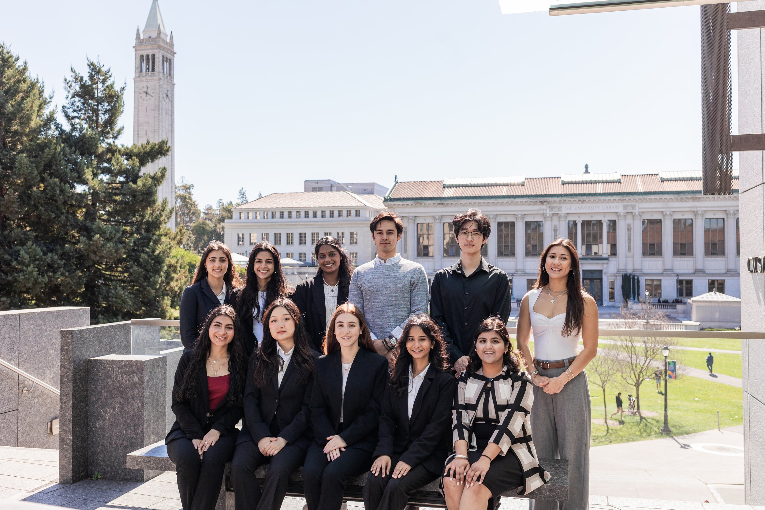 Group of young adults, girls and boys, posing outdoors in front of a university building with a clock tower, trees, and a bright clear sky.