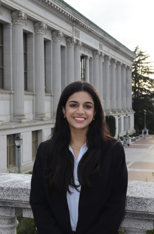 A young woman with long black hair smiling in front of a historic building with large columns, during late afternoon or early evening.
