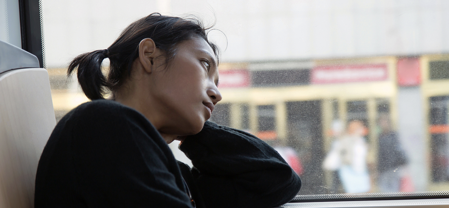 A woman sitting on a bus or train, looking out the window with a thoughtful or contemplative expression.