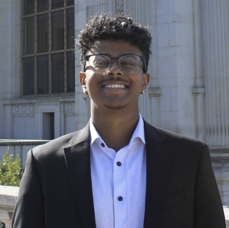 Young man with curly hair, glasses, and a suit smiling outdoors in front of a historic building.