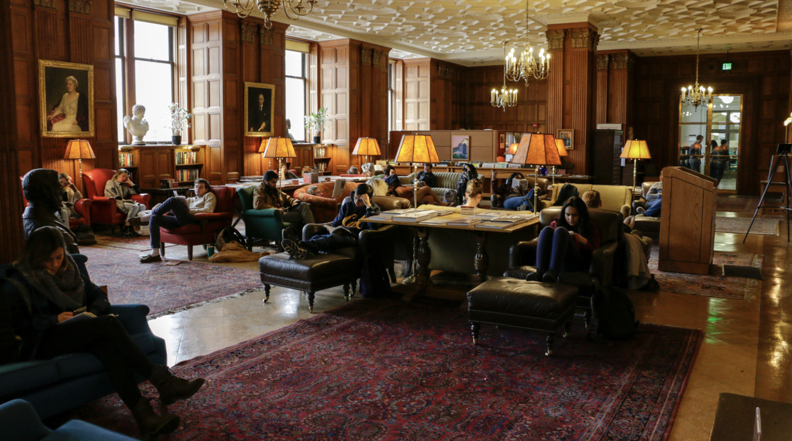 A spacious library lounge with wooden paneled walls, multiple windows, and several people sitting on chairs and sofas, some reading or using devices. There are table lamps, framed portraits, and bookshelves.