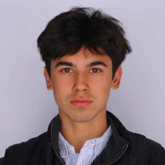 Portrait of a young man with dark hair, wearing a light blue collared shirt and a black jacket, against a plain gray background.
