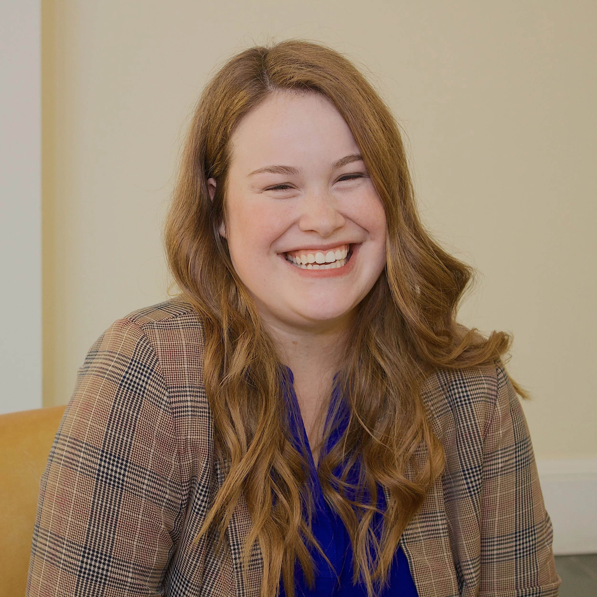 A young woman with long wavy red hair, wearing a brown checkered blazer and a blue top, smiling openly in an indoor setting with a plain beige wall in the background.
