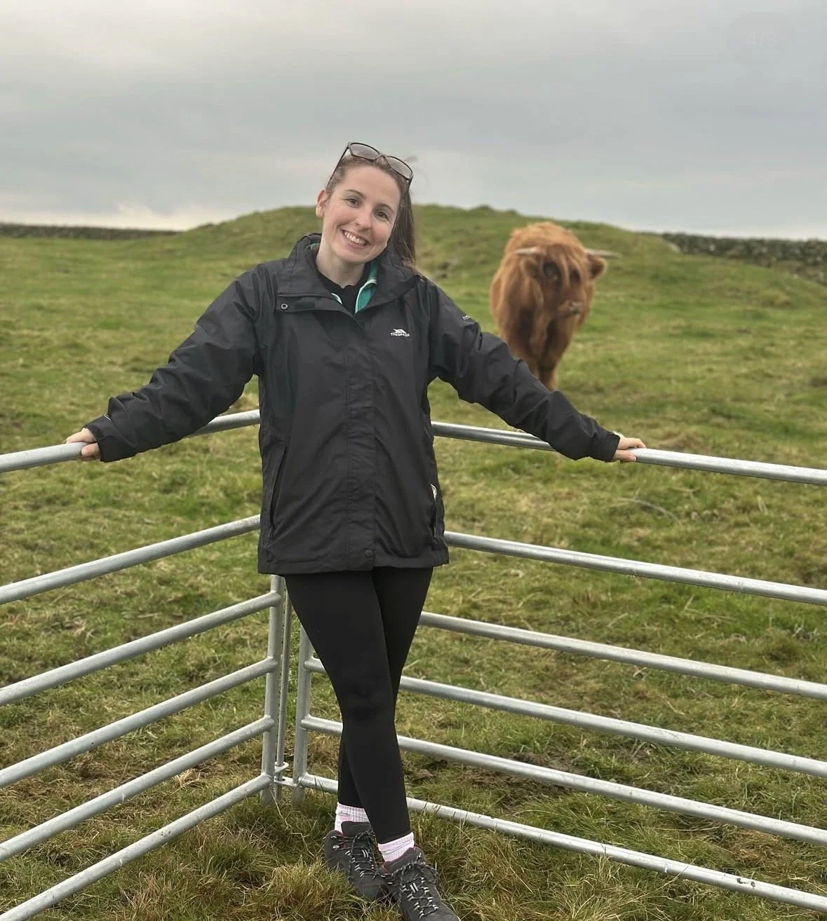 A smiling woman in a black jacket and black leggings standing on a grassy field with a brown cow behind a metal fence on a cloudy day.